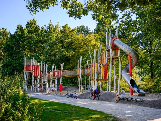 Moderner Abenteuerspielplatz mit Rutschen, Klettergerüsten und Besuchern umgeben von grüner Natur.