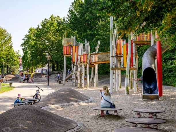 Wasserspielplatz Emssee Wiedenbrück 2 Moderner Spielplatz mit bunten Klettergerüsten, Rutsche und Sandfläche, umgeben von grünen Bäumen.
