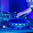 Ein DJ legt im Nachtclub Platten auf, während bunte Lichter die Mischpulte beleuchten.