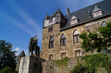 Schloss Burg Ritterstatue vor historischem Schlossgebäude aus Naturstein unter blauem Himmel, umgeben von Grün.