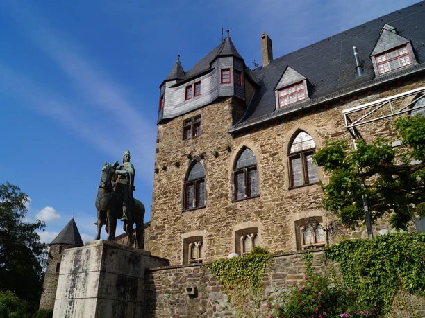 Schloss Burg Ritterstatue vor historischem Schlossgebäude aus Naturstein unter blauem Himmel, umgeben von Grün.