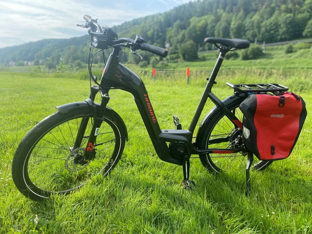Černé elektrokolo Haibike s červenou brašnou stojí za slunečného počasí na zelené louce před zalesněnou kopcovitou krajinou.