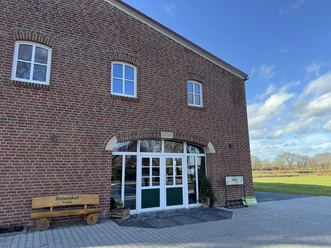Biolandhof Freese Verkaufsladen, Gemeinde Rhauderfehn Eingang eines Backsteingebäudes mit grünen Türen, daneben eine Bank mit der Aufschrift Biolandhof Freese und ein Schild, umgeben von einer gepflasterten Fläche und Wiesen unter blauem Himmel