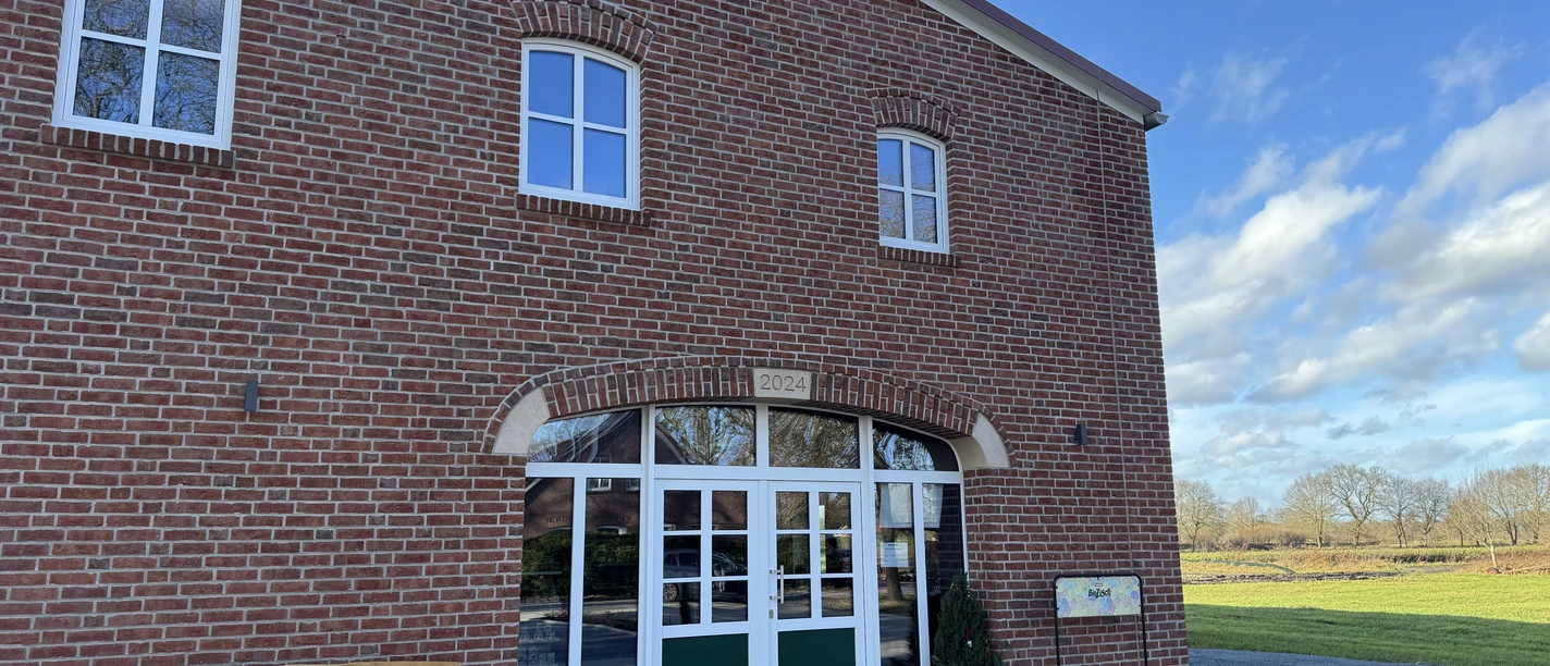 Biolandhof Freese Verkaufsladen, Gemeinde Rhauderfehn Eingang eines Backsteingebäudes mit grünen Türen, daneben eine Bank mit der Aufschrift Biolandhof Freese und ein Schild, umgeben von einer gepflasterten Fläche und Wiesen unter blauem Himmel