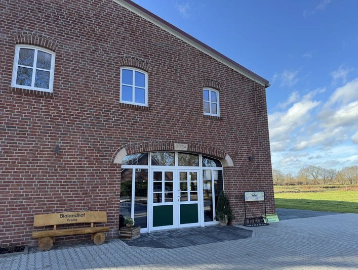 Biolandhof Freese Verkaufsladen, Gemeinde Rhauderfehn Eingang eines Backsteingebäudes mit grünen Türen, daneben eine Bank mit der Aufschrift Biolandhof Freese und ein Schild, umgeben von einer gepflasterten Fläche und Wiesen unter blauem Himmel