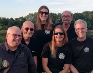 New Foggy Few Sechs lächelnde Personen tragen Sonnenbrillen, stehen dicht beieinander vor einer grünen Landschaft.