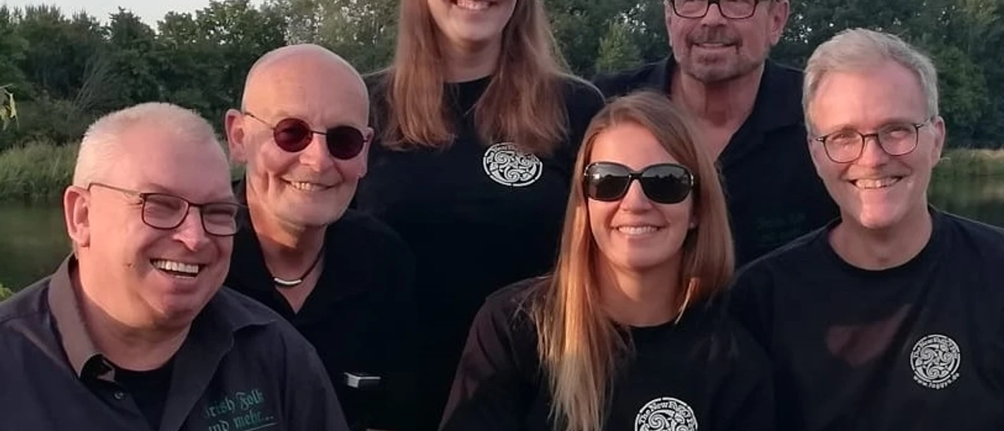 New Foggy Few Sechs lächelnde Personen tragen Sonnenbrillen, stehen dicht beieinander vor einer grünen Landschaft.