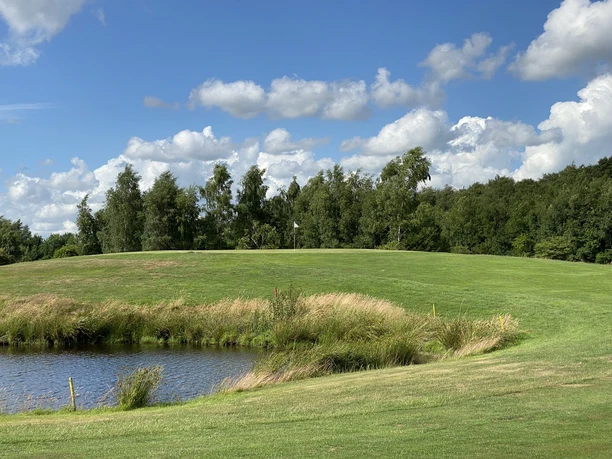 Blauer Fasan Golf 1.jpg Eine gepflegte, grüne Golfanlage mit einem Teich im Vordergrund. Am Horizont stehen Bäume unter einem blauen Himmel mit weißen Wolken.
