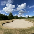 Blauer Fasan Golf 4.jpg Harmonisch geschwungener Sandbunker auf grünem Golfplatz unter blauem Himmel mit weißen Wolken.