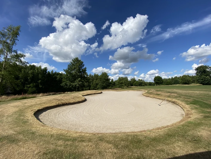 Blauer Fasan Golf 4.jpg Harmonisch geschwungener Sandbunker auf grünem Golfplatz unter blauem Himmel mit weißen Wolken.