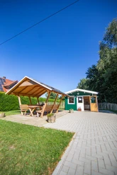 In't Euwer Holzhütte mit überdachter Sitzgruppe auf gepflastertem Platz neben grüner Hecke unter blauem Himmel.