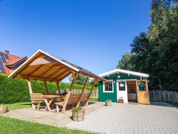 In't Euwer Holzhütte mit überdachter Sitzgruppe auf gepflastertem Platz neben grüner Hecke unter blauem Himmel.