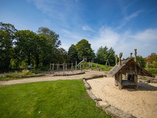 C5D_7050_MedienAtleierEmsland.jpg Holzspielplatz mit Rutschen, Schaukeln und Sandfläche in grüner Parklandschaft unter blauem Himmel