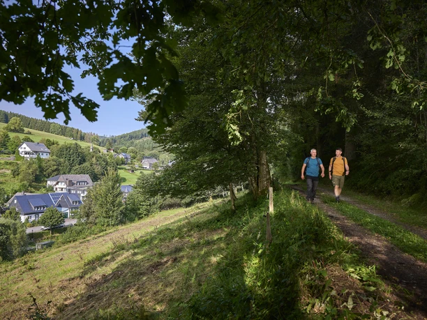 Zwei Wanderer im grünen Wald mit Ausblick auf den Ort Latrop am Rothaarsteig Zwei Wanderer im grünen Wald mit Ausblick auf den Ort Latrop am Rothaarsteig