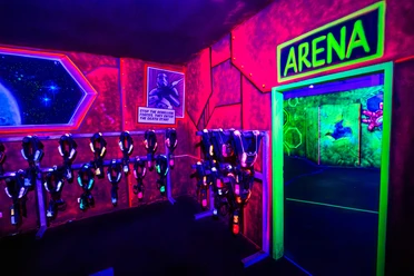 Laser Taq Arena Heros.jpg Neonbeleuchteter Laserspielplatz mit futuristischem Raumdekor und leuchtenden Laserwaffen an der Wand.