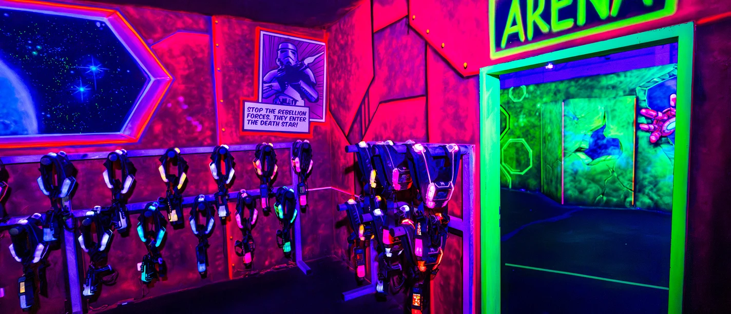 Laser Taq Arena Heros.jpg Neonbeleuchteter Laserspielplatz mit futuristischem Raumdekor und leuchtenden Laserwaffen an der Wand.