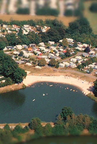 Luftaufnahme eines großen, von Wald umgebenen Campingplatzes mit Wohnwagen und Zelten an einem See.