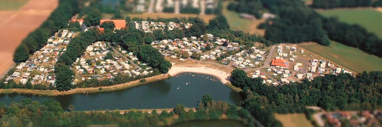 Luftaufnahme.jpg Luftaufnahme eines großen, von Wald umgebenen Campingplatzes mit Wohnwagen und Zelten an einem See.