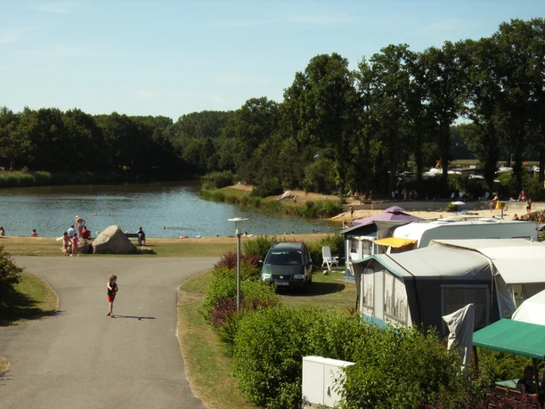 Campingplatz am See mit Wohnwagen, Badegästen am Sandstrand und bewaldeter Umgebung im Sommer.