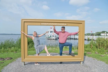 paar-im-bilderrahmen-vor-zwischenahner-meer.jpg Fröhliches Paar posiert im Holz-Bilderrahmen am Zwischenahner Meer, dahinter ein Schiff.