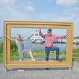 paar-im-bilderrahmen-vor-zwischenahner-meer.jpg Fröhliches Paar posiert im Holz-Bilderrahmen am Zwischenahner Meer, dahinter ein Schiff.