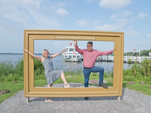 paar-im-bilderrahmen-vor-zwischenahner-meer.jpg Fröhliches Paar posiert im Holz-Bilderrahmen am Zwischenahner Meer, dahinter ein Schiff.