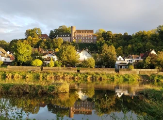 Burg Herstelle thront auf einem Hügel, umgeben von grünen Bäumen und spiegelt sich im Fluss darunter.