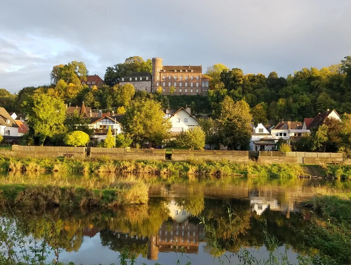 Burg Herstelle thront auf einem Hügel, umgeben von grünen Bäumen und spiegelt sich im Fluss darunter.