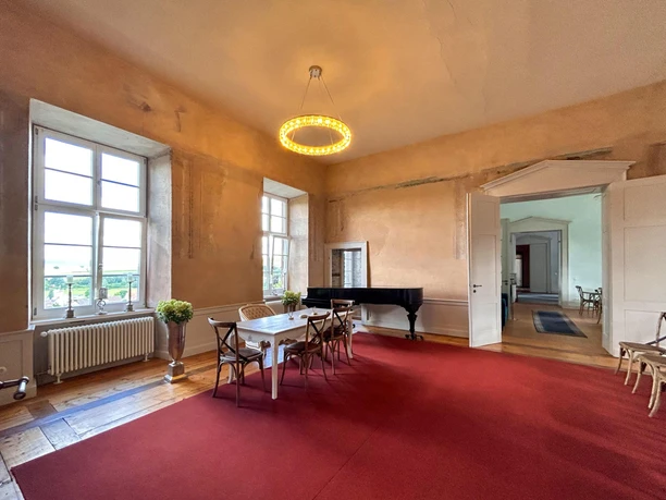 Heller Saal mit rotem Teppich, stilvollen Stühlen um einen Tisch und einem schwarzen Flügel.