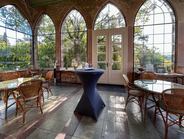 Wintergarten der Burg Herstelle mit großen Glasfenstern, eleganten Rattanstühlen und Tischen.