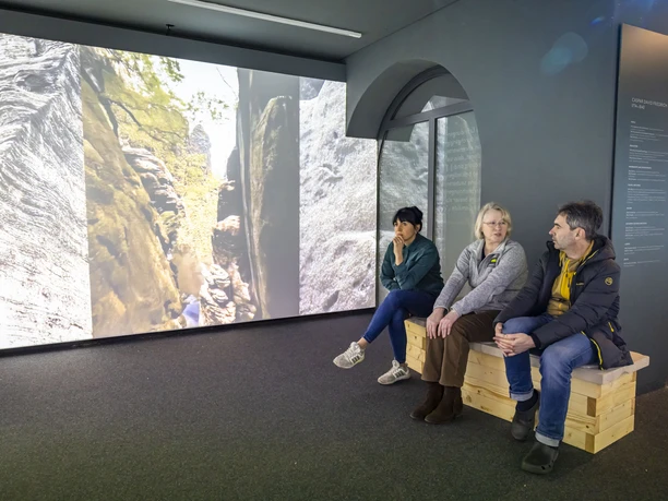 Tři lidé sedí na dřevěné lavičce v muzeu a dívají se na velkou projekci skal a stromů. Atmosféra je klidná a zamyšlená.