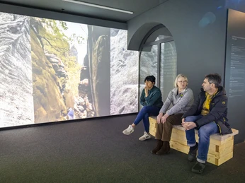 Mediale Ausstellung „CDFriedrich inspiriert“ Drei Personen sitzen auf einer Holzbank in einem Museum, betrachten eine große Projektion von Felsen und Bäumen. Die Atmosphäre ist ruhig und nachdenklich.Three people are sitting on a wooden bench in a museum, looking at a large projection of rocks and trees. The atmosphere is calm and thoughtful.Tři lidé sedí na dřevěné lavičce v muzeu a dívají se na velkou projekci skal a stromů. Atmosféra je klidná a zamyšlená.Trzy osoby siedzą na drewnianej ławce w muzeum, patrząc na dużą projekcję skał i drzew. Atmosfera jest spokojna i pełna zadumy.Drie mensen zitten op een houten bank in een museum en kijken naar een grote projectie van rotsen en bomen. De sfeer is kalm en bedachtzaam.Tre persone sono sedute su una panchina di legno in un museo e guardano una grande proiezione di rocce e alberi. L'atmosfera è calma e riflessiva.