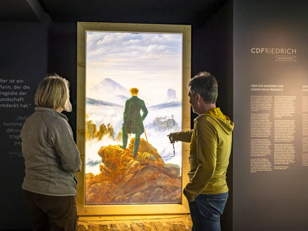 Mediale Ausstellung „CDFriedrich inspiriert“ Zwei Personen betrachten ein beleuchtetes Gemälde von Caspar David Friedrich in einer Ausstellung, umgeben von erklärenden Texten an der Wand.