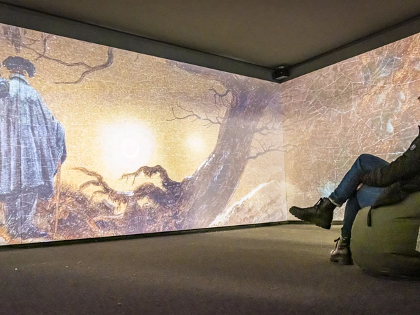 Mediale Ausstellung "CDFriedrich inspiriert“ Osoba sedí na pytli s fazolemi v místnosti s velkou projekcí obrazu, na němž jsou dva muži v kabátech.