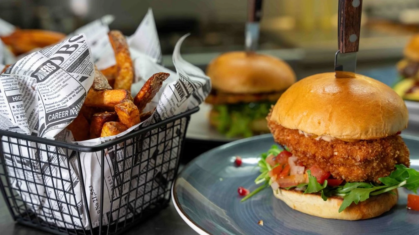 Max Burger & Bar Ein knuspriger Hähnchenburger auf einem blauen Teller neben einem Korb mit würzigen Pommes frites.