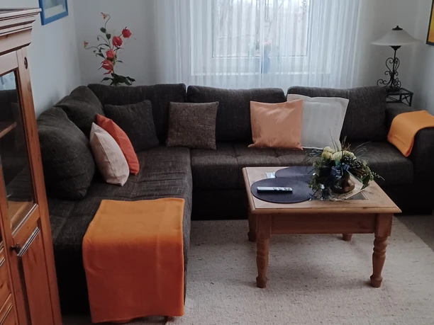 Gästewohnung Werkerwald-Wohnzimmer-c-Dieter Westermann.jpg Wohnzimmer mit braunem Ecksofa, orangen Decken, Holztisch mit Blumengesteck und hellem Teppich.