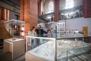 meissen-c-philipp-herfort-photography-4409.jpg Ein Paar betrachtet ein detailliertes Modell in einem lichtdurchfluteten Museum mit hohen Decken.