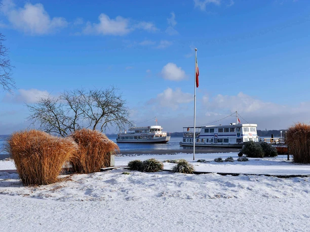 kurpark-im-winter-weisse-flotte.jpg Verschneiter Kurpark in Bad Zwischenahn vor blauem Himmel. Zwei Fahrgastschiffe der Weißen Flotte am Steg.