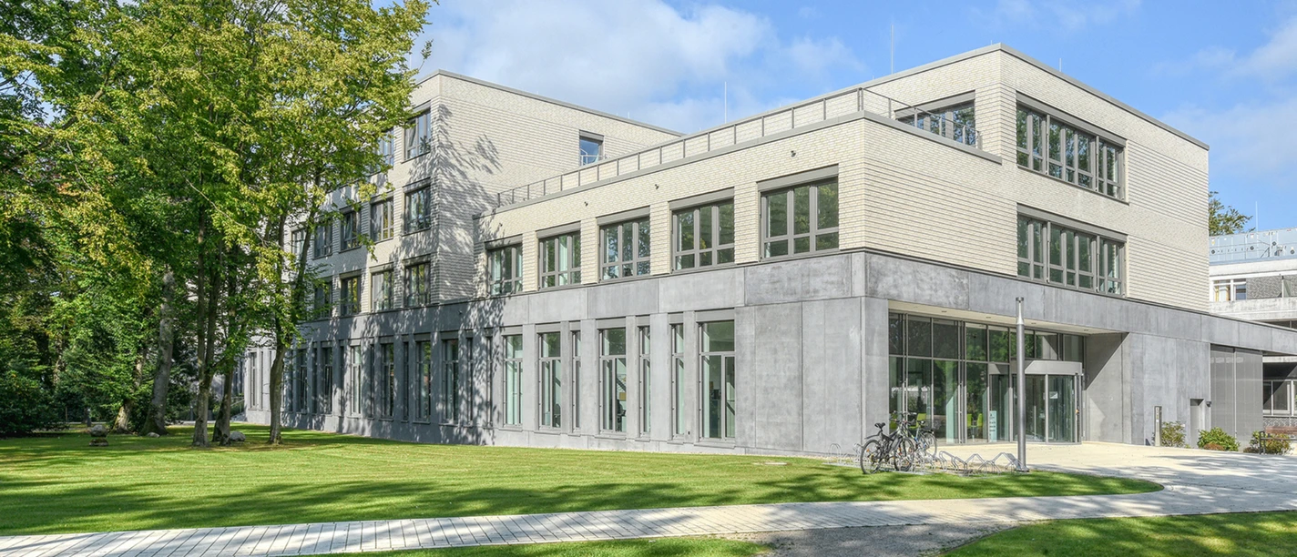 reha-zentrum-am-meer.png Das Reha-Zentrum am Meer ist ein modernes Gebäude mit hellem Backstein und großen Fenstern, umgeben von Grünflächen und Bäumen. Fahrräder stehen vor dem Eingang, was eine einladende und ruhige Atmosphäre schafft.