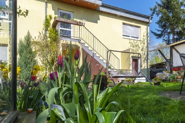 Eingang Hinterhofgarten mit blühenden Tulpen im Vordergrund vor einem gelben Haus mit Außentreppe.