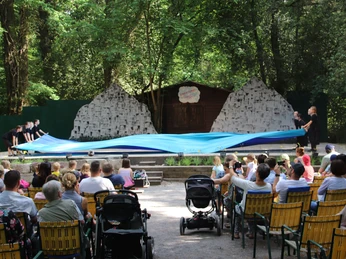 2018-Jim-Knopf-und-Lukas-der-Lokomotivführer-1-Waldbühne-Otternhagen Besucher sitzen auf einer Freilichtbühne im Wald, während Darsteller ein großes blaues Tuch bewegen.