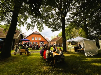 Sommerfest 2024 im Museumsgarten des Fehn- und Schiffahrtsmuseums Westrhauderfehn IMG_9146.JPG Menschen sitzen an Tischen und Bänken im Garten vor einem roten Backsteinhaus unter Bäumen bei einer Veranstaltung mit Bühne und Pavillons