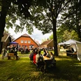 Sommerfest 2024 im Museumsgarten des Fehn- und Schiffahrtsmuseums Westrhauderfehn IMG_9146.JPG Menschen sitzen an Tischen und Bänken im Garten vor einem roten Backsteinhaus unter Bäumen bei einer Veranstaltung mit Bühne und Pavillons