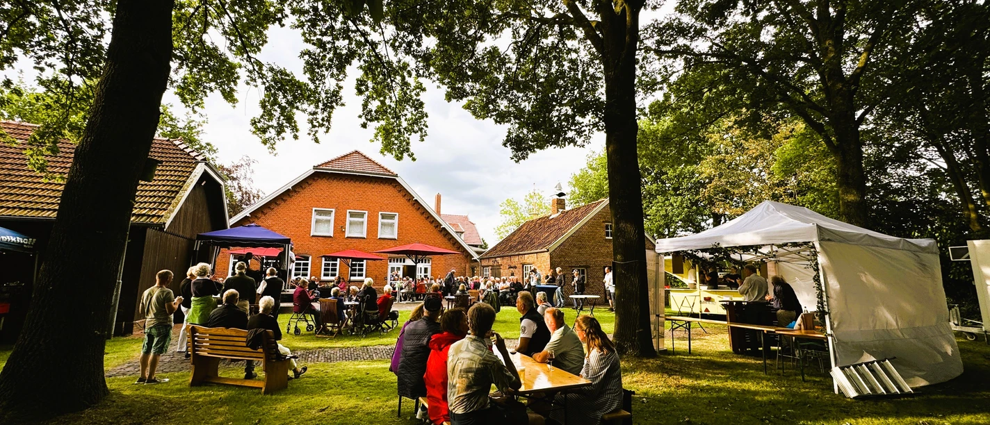Sommerfest 2024 im Museumsgarten des Fehn- und Schiffahrtsmuseums Westrhauderfehn IMG_9146.JPG Menschen sitzen an Tischen und Bänken im Garten vor einem roten Backsteinhaus unter Bäumen bei einer Veranstaltung mit Bühne und Pavillons