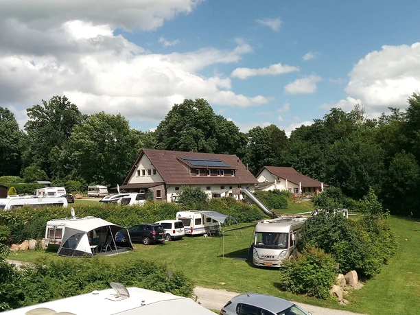 Ein Campingplatz mit Wohnmobilen und Zelten in grüner Umgebung, von Bäumen umgeben und sonnig.