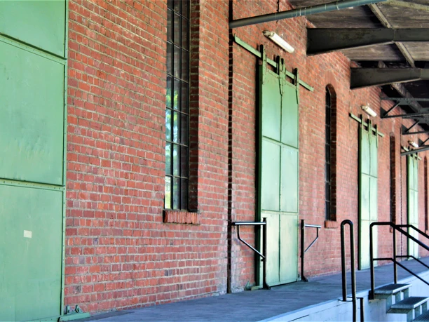 Roter Backsteinbau des ehemaligen Güterbahnhofs mit grünen Türen und Fenstern, überdachter Bereich.
