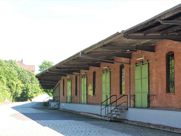 Backsteingebäude mit grünen Türen und Vordächern, Kopfsteinpflasterweg, Grünfläche an der Seite.