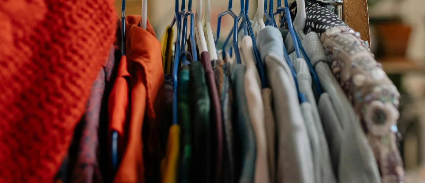 Second Hand (c) pexels Eine hand greift nach einem Kleiderständer, an dem viele verschiedene Kleidungsstücke an Kleiderbügeln hängen.
