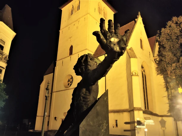 Herford-Schauergeschichten-quer -Pro-Herford-GmbH.jpg Eine imposante Skulptur vor einer beleuchteten, gotischen Kirche bei Nacht, Herford.
