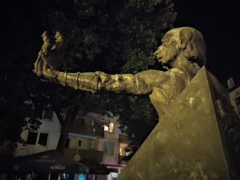 Herford-Schauergeschichten-Pro-Herford-GmbH.jpg Bronzestatue eines Mannes mit erhobenem Arm, in der Nacht beleuchtet, vor Bäumen und Gebäuden.